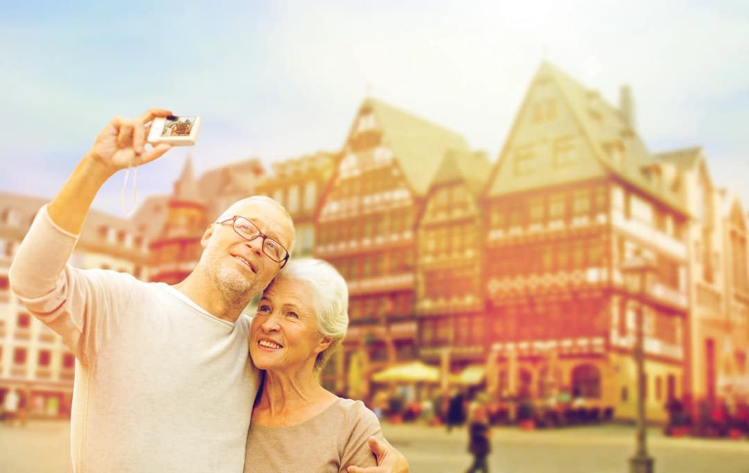 A happy senior couple taking a selfie with a compact camera, smiling in front of a blurred background of historic half-timbered buildings in a European town square.