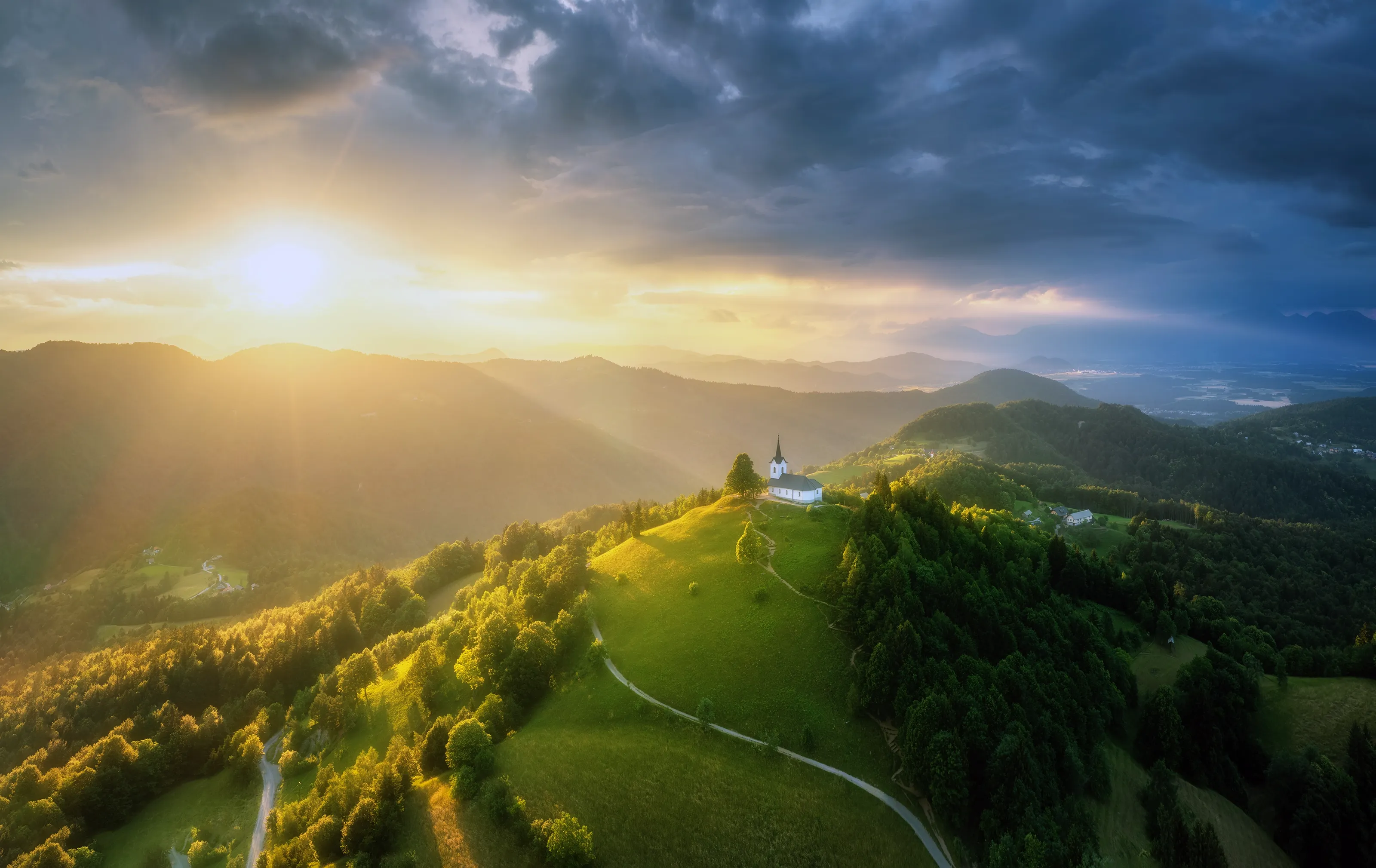 A stunning aerial view of a picturesque church on a green hilltop in Slovenia, bathed in the golden rays of a dramatic sunset with mountains in the background.